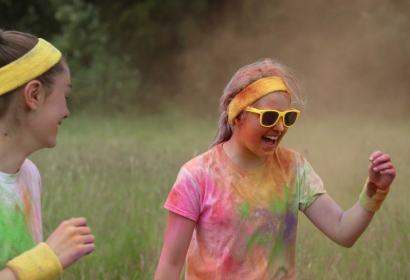 a girl covered in coloured powder wearing yellow sunglasses and headband laughs happily